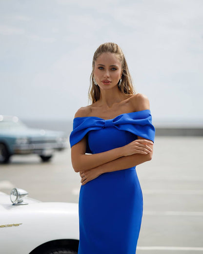 Woman in a blue off-shoulder dress standing in front of vintage cars.