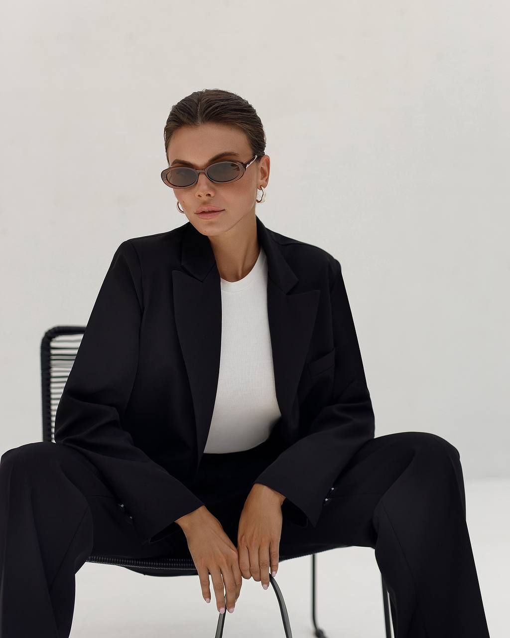 Person wearing a black suit and white shirt sitting on a chair against a plain background