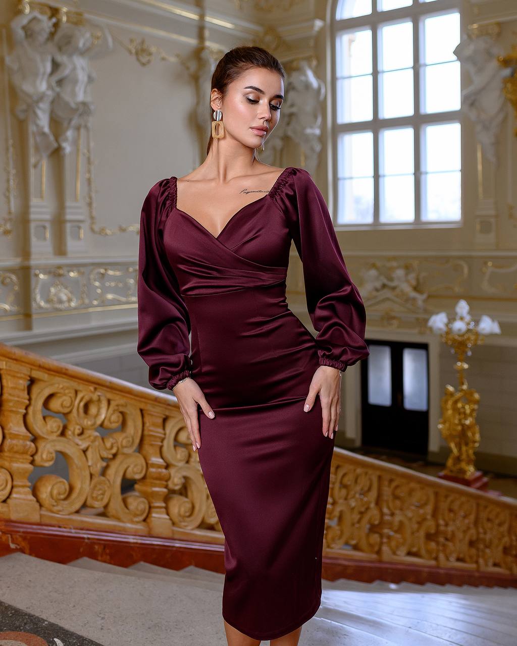 Woman in a burgundy dress standing in an ornate room with decorative walls and a large window.