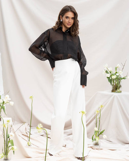 Woman wearing a black sheer blouse and white pants standing in a minimalistic setting with flowers.