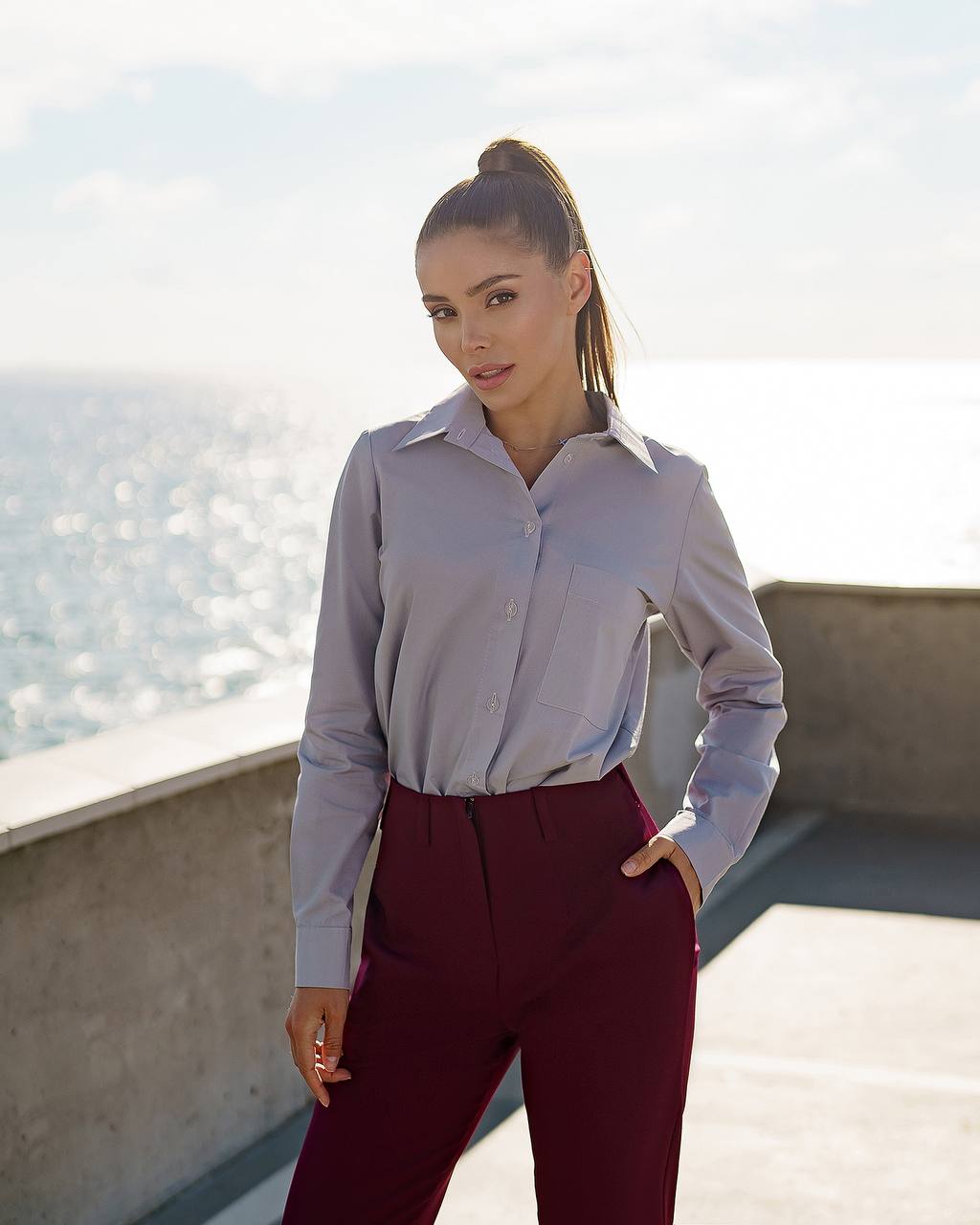 Woman wearing a light gray shirt and maroon pants standing on a rooftop with a cityscape in the background.