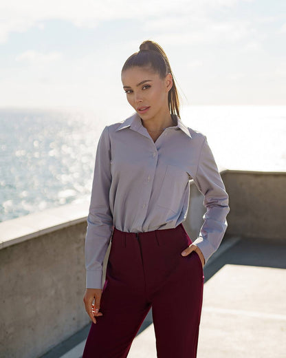 Woman wearing a light gray shirt and maroon pants standing on a rooftop with a cityscape in the background.
