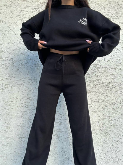 Person wearing a black oversized sweatshirt and pants against a textured wall.