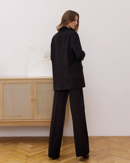 Woman wearing a black suit standing in a room with wooden flooring and a light-colored wall.