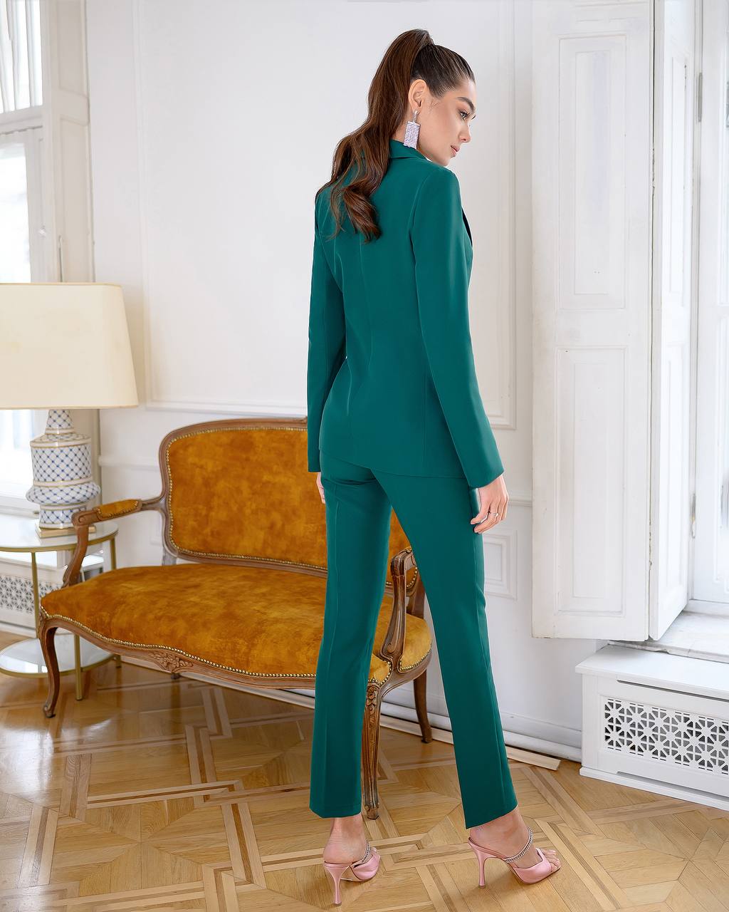 Woman in a teal suit standing in a room with a vintage chair and lamp.