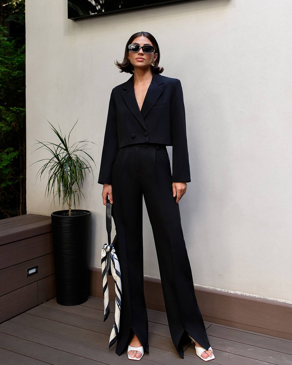 Woman in a black suit standing outdoors with a plant and wooden bench in the background
