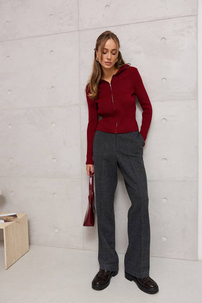 Woman wearing a red sweater and gray pants against a light gray wall.