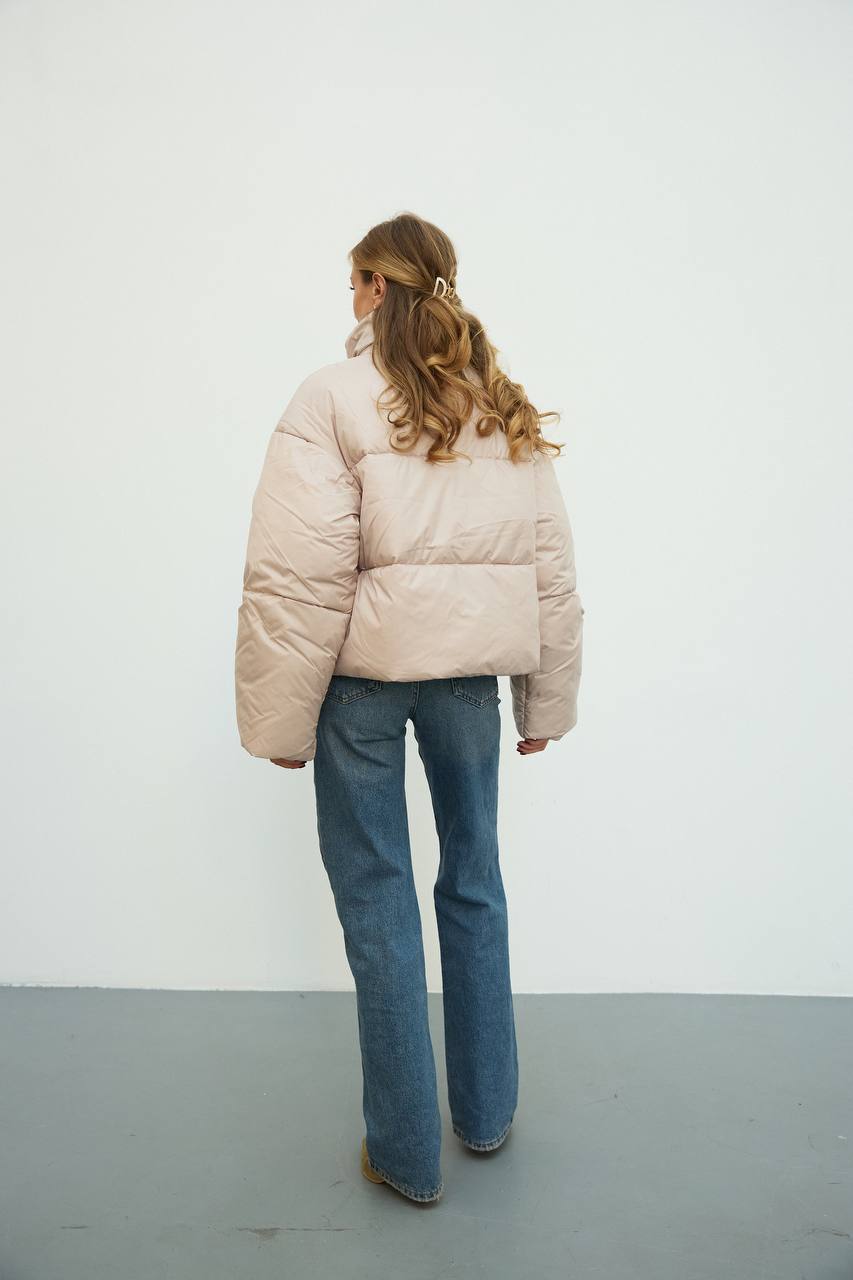 Person wearing a beige puffer jacket and blue jeans against a white wall.