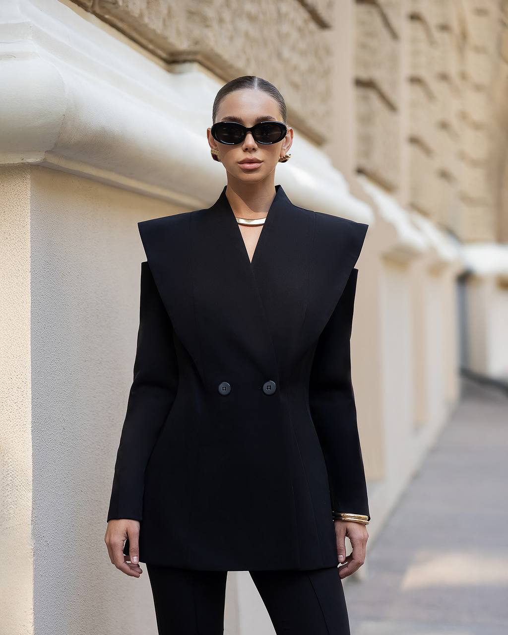 A model wearing a black double-breasted blazer with dramatic shoulder flaps and structured shoulders, paired with matching black trousers, black sunglasses, and gold jewelry, standing outdoors against a beige stone building.