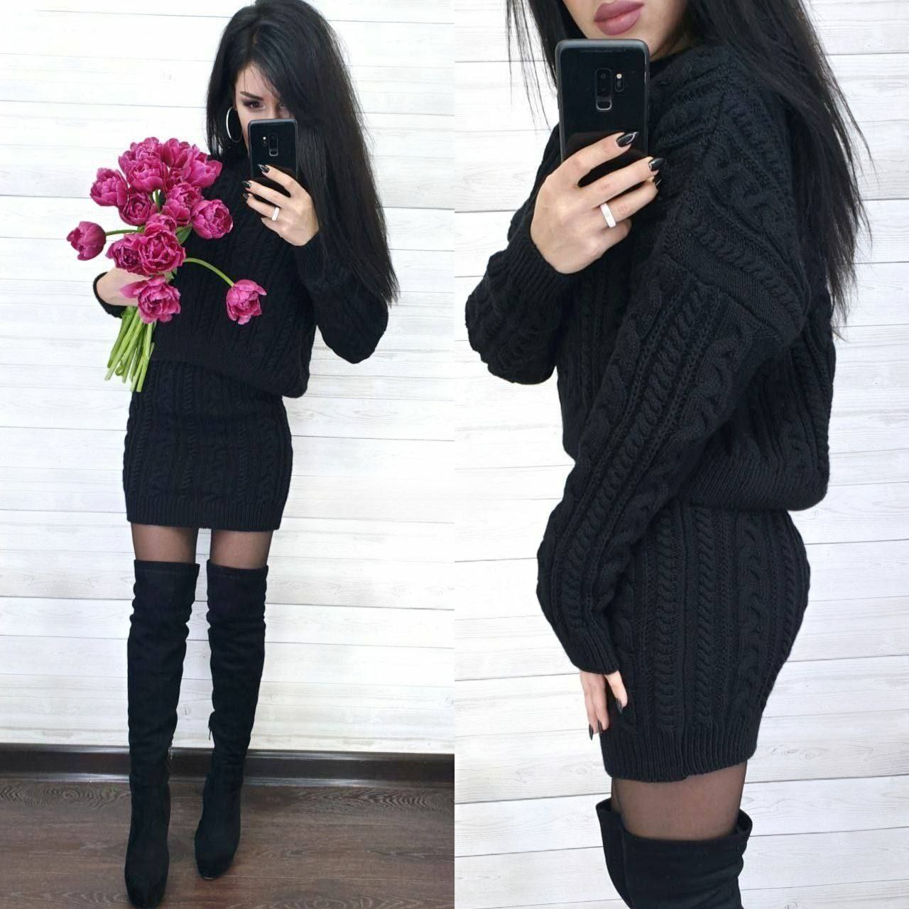 Woman wearing a black cable knit sweater dress with knee-high boots, holding pink flowers.