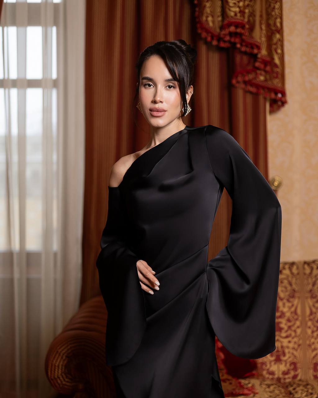 A woman wearing a black satin one-shoulder dress with dramatic draped sleeves, standing in an elegant room with brown curtains and patterned upholstery.