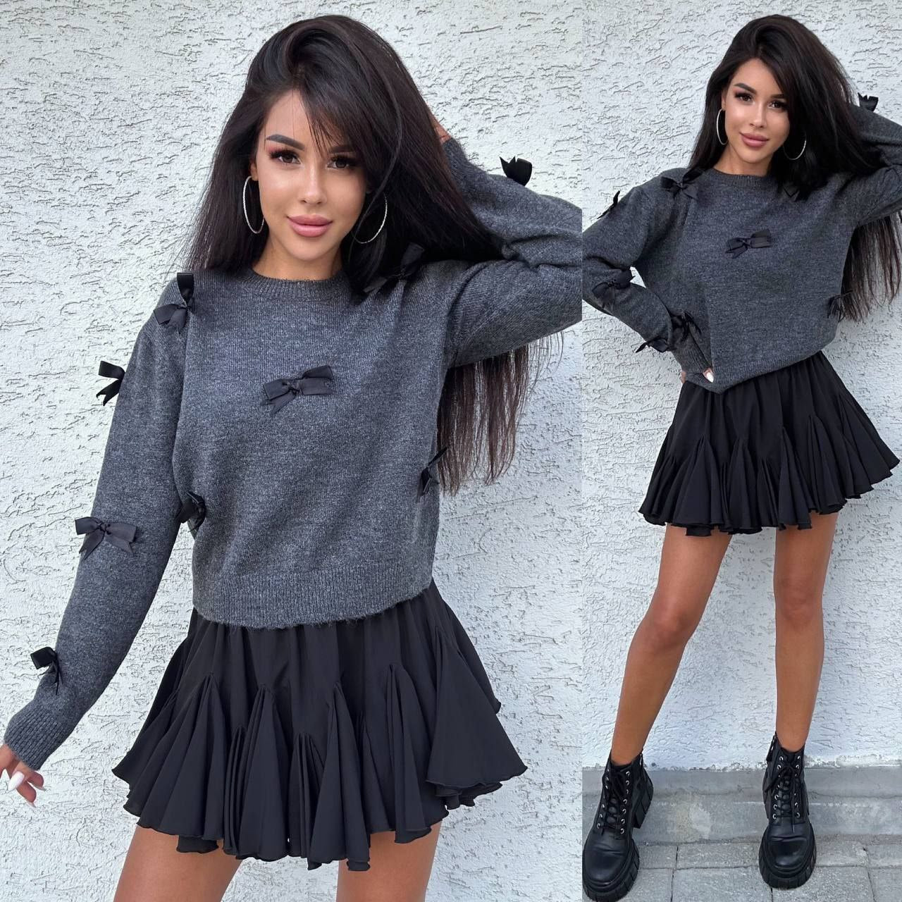 Woman wearing a gray sweater with black bows and a black skirt against a light gray wall.