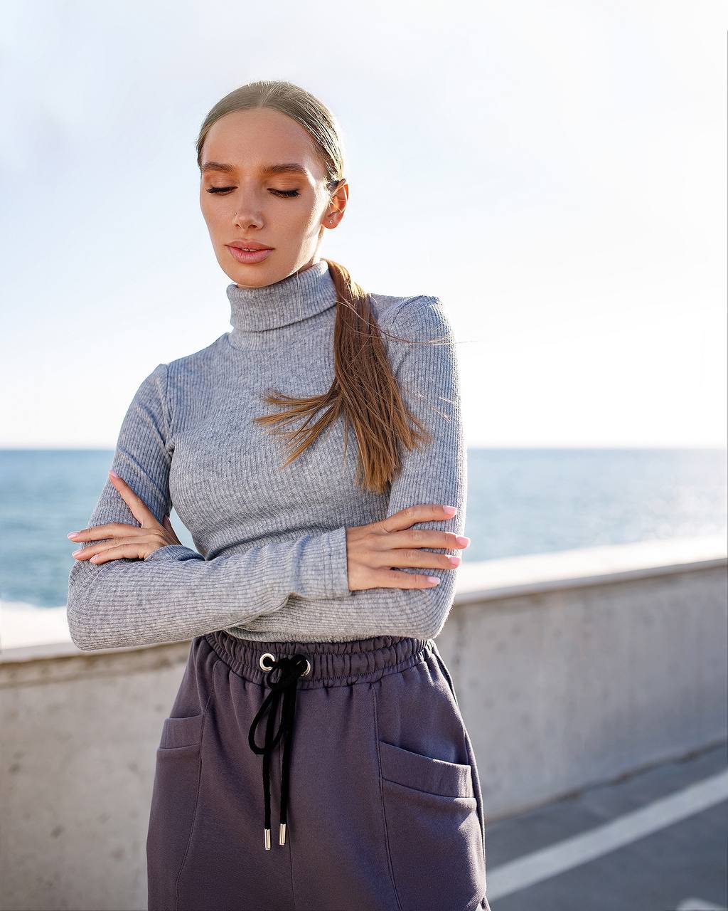 Woman wearing a gray turtleneck sweater and purple pants standing by a coastal area.