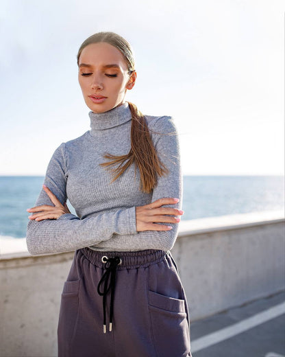 Woman wearing a gray turtleneck sweater and purple pants standing by a coastal area.