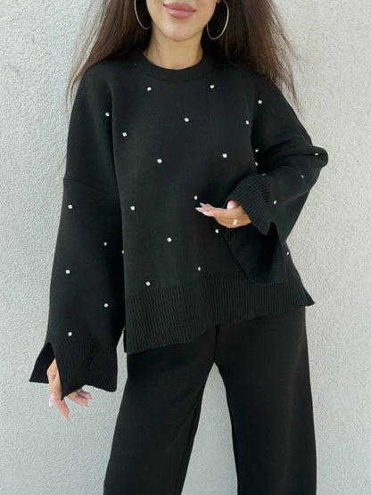 Black sweater with pearl embellishments worn by a person against a gray background