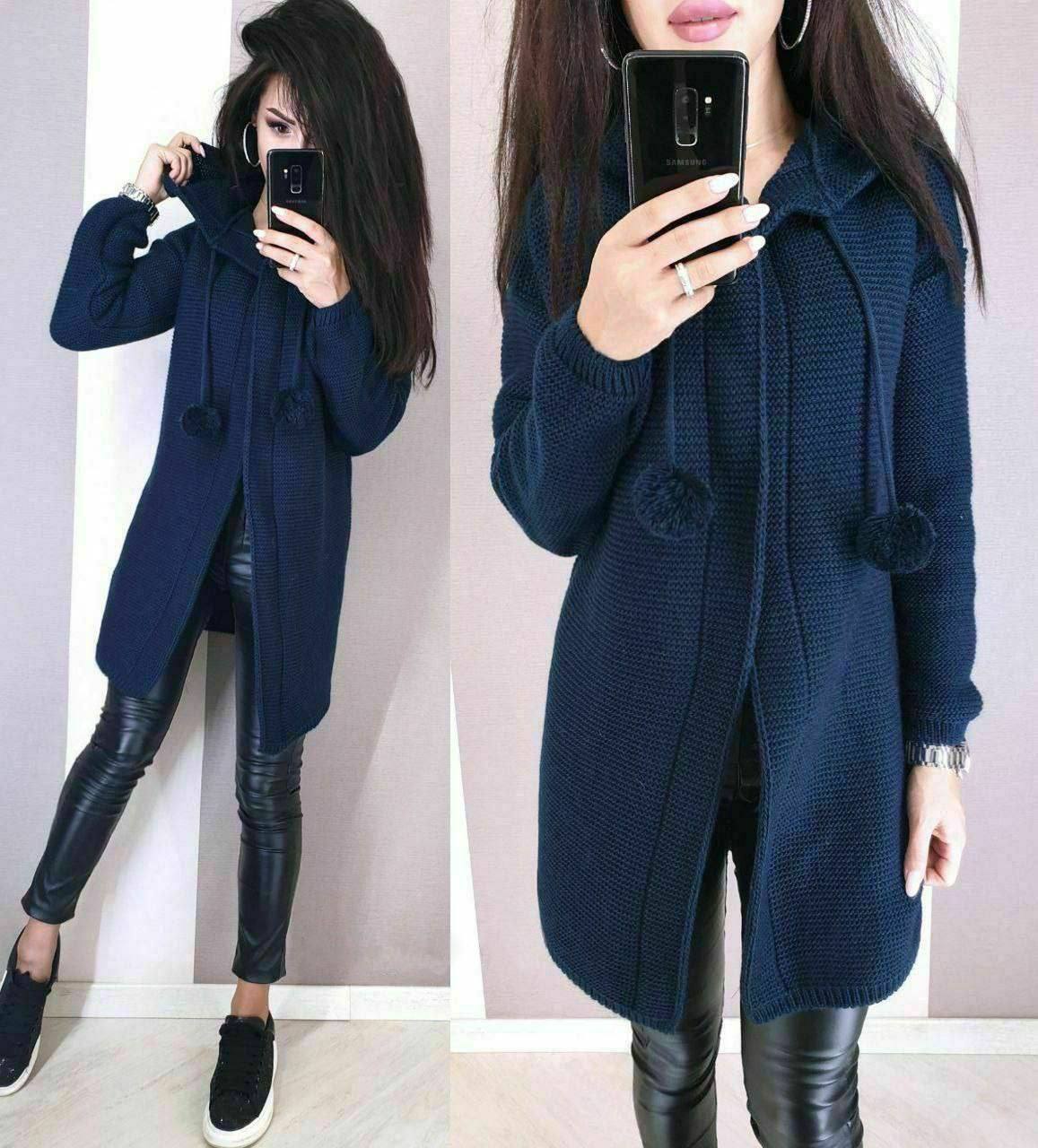 Woman wearing a navy blue knitted cardigan with pom-poms, standing against a plain background.