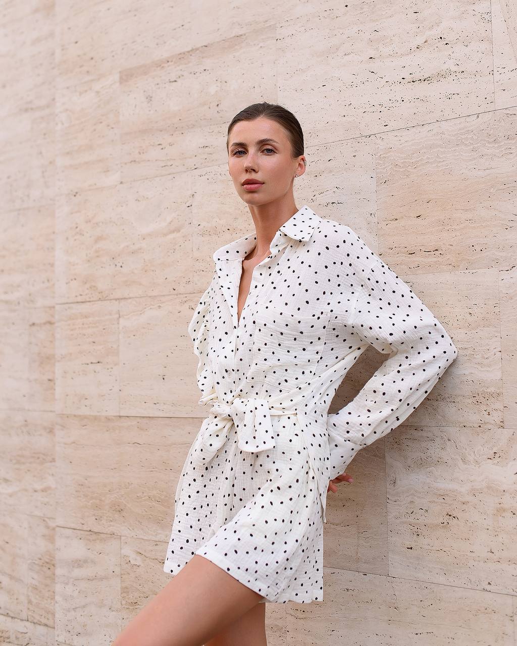 Woman wearing a white polka dot dress against a beige stone wall