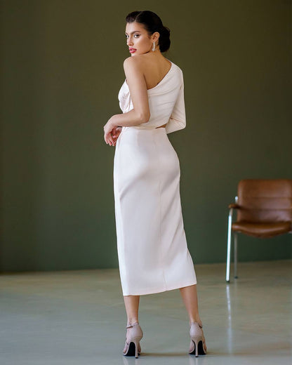 Woman in a white one-shoulder dress standing in a room with a brown chair.