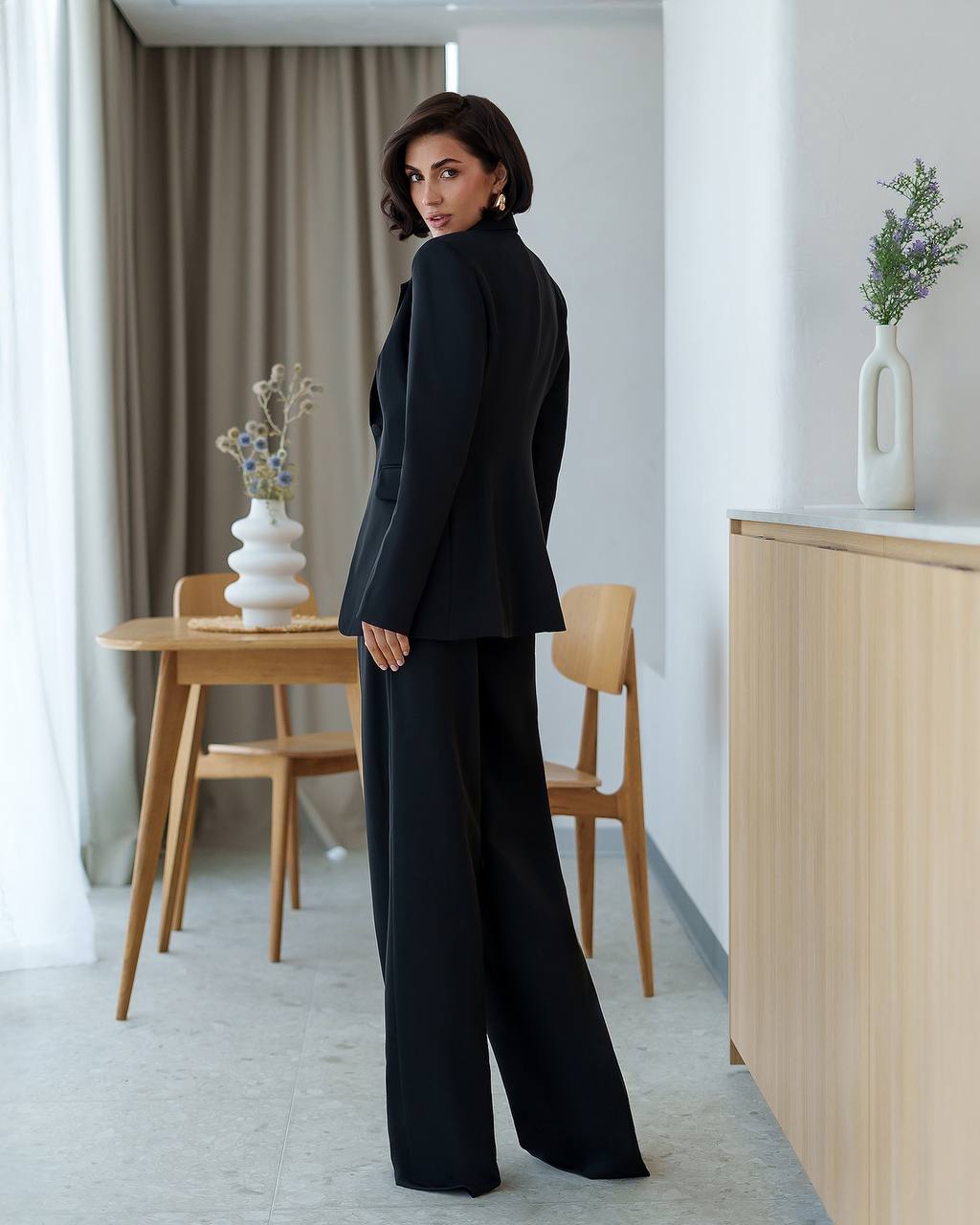 Woman in a black suit standing in a modern interior setting with wooden furniture and decorative plants.