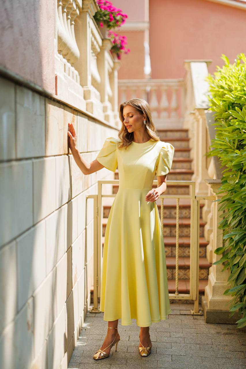 A woman wearing a pale yellow midi dress with puff sleeves and gold heels, posing on a stone walkway next to a building wall with a staircase in the background.