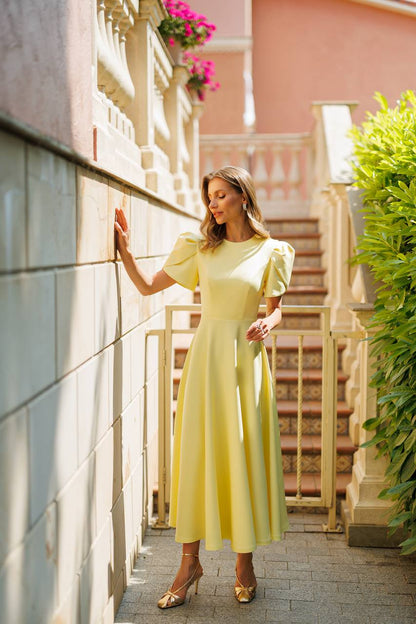 A woman wearing a pale yellow midi dress with puff sleeves and gold heels, posing on a stone walkway next to a building wall with a staircase in the background.