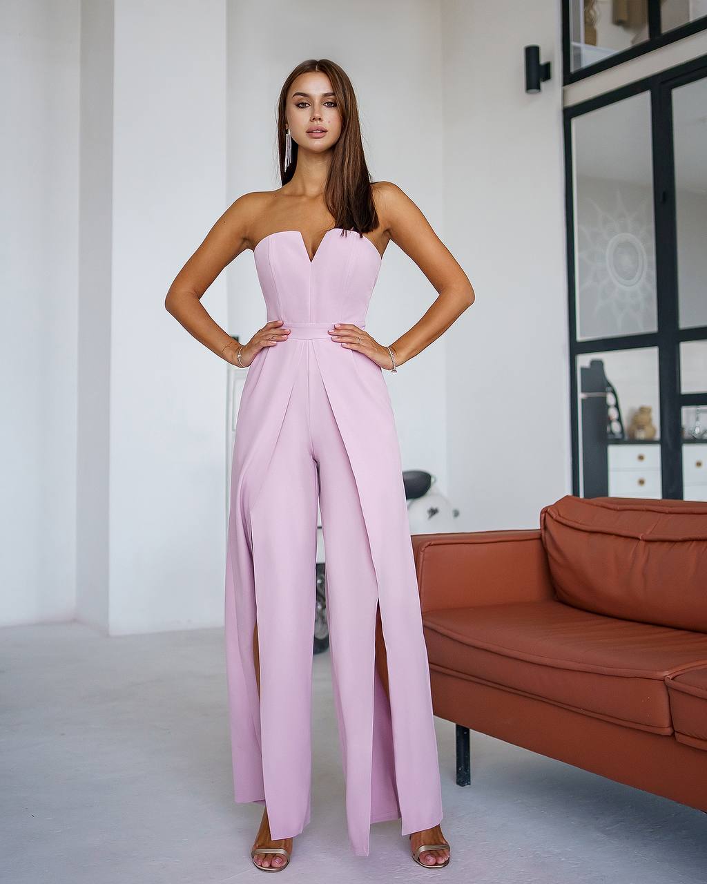 Woman wearing a pink strapless jumpsuit in a room with a brown sofa.
