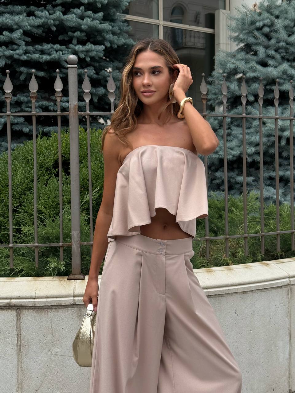 Woman wearing a beige strapless top and wide-leg pants standing outdoors.