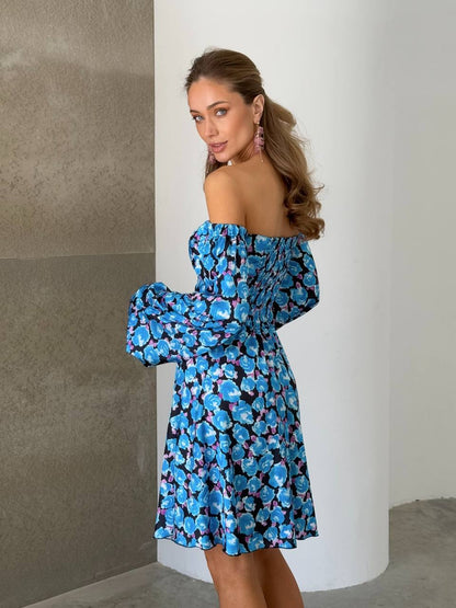 Woman wearing a blue floral off-shoulder dress against a neutral wall.