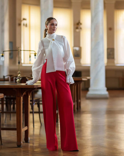 Woman in a white blouse and red pants standing in an elegant interior setting.