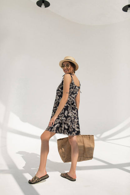 Woman in a black floral dress with a straw hat and bag, standing against a white background
