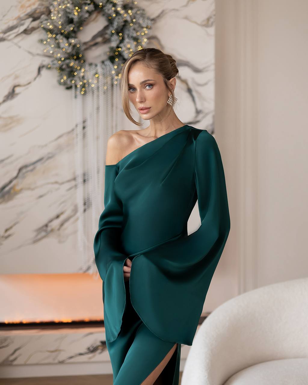 A woman models a dark teal green one-shoulder evening gown with dramatic draped sleeves in an elegant indoor setting with a marble wall and fireplace.