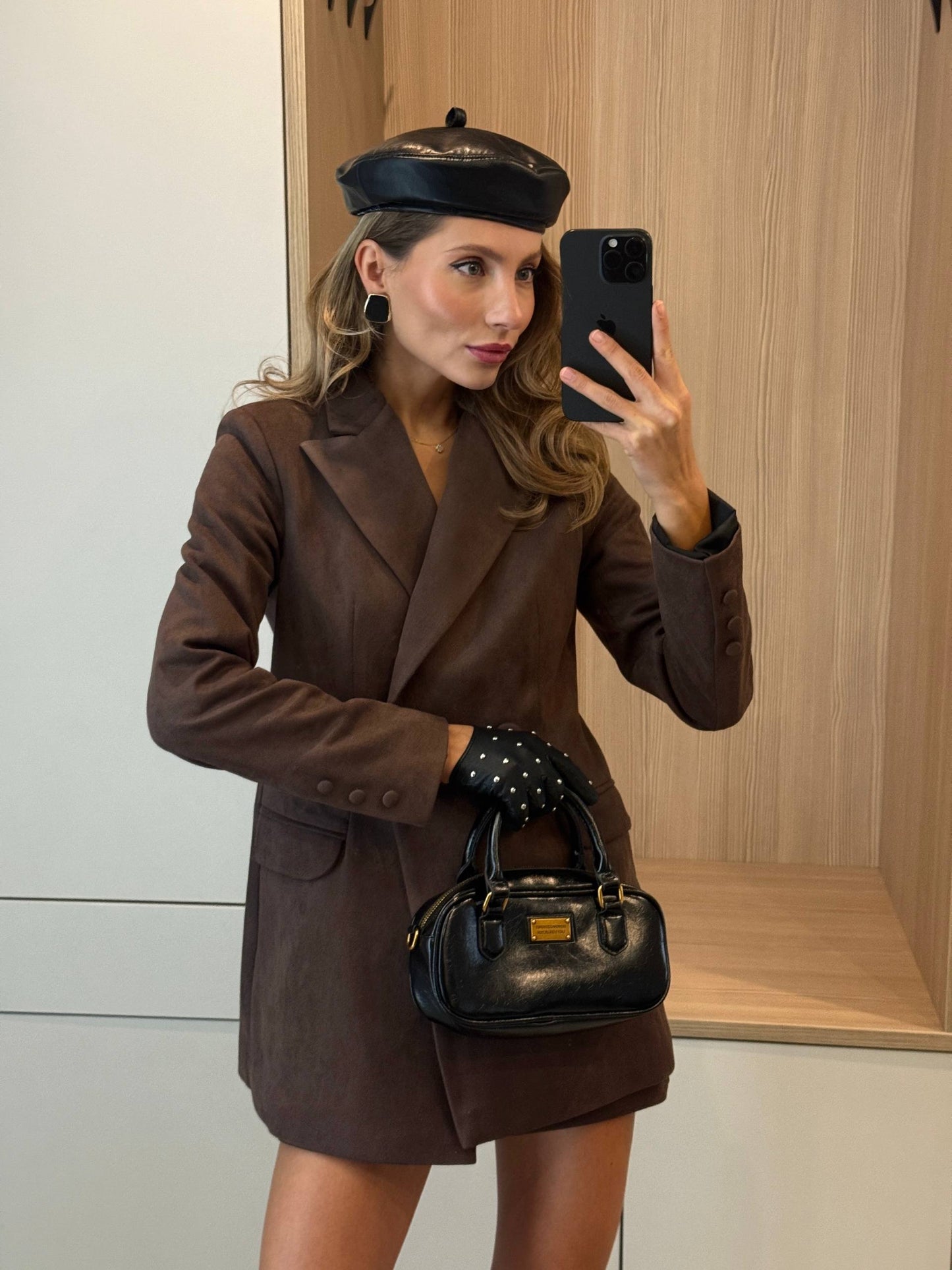 A woman takes a mirror selfie wearing a brown double-breasted blazer, a matching brown mini skirt, a black beret, black studded gloves, and carrying a black leather handbag.