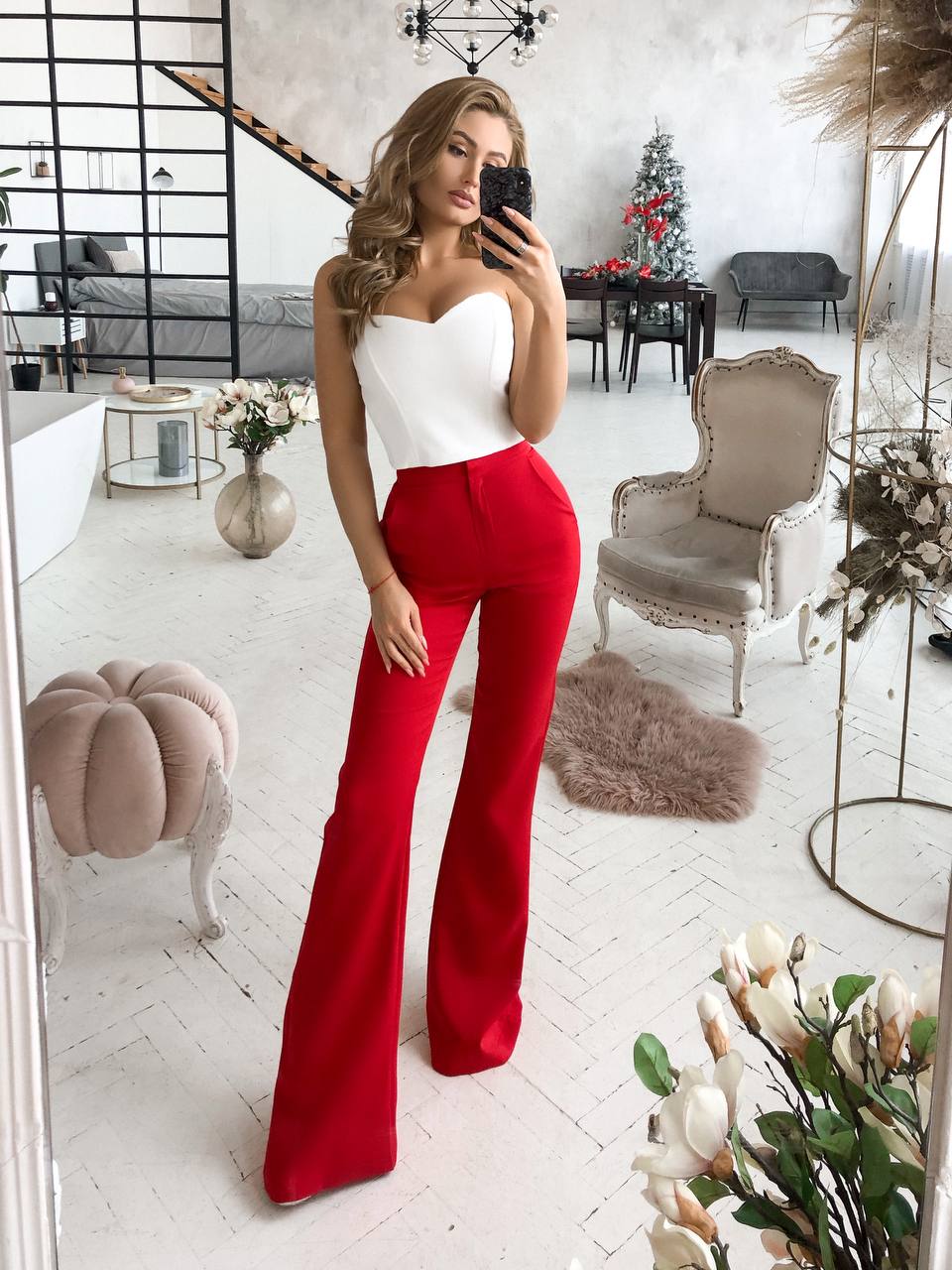Woman taking a mirror selfie wearing a white top and red pants in a stylish room.