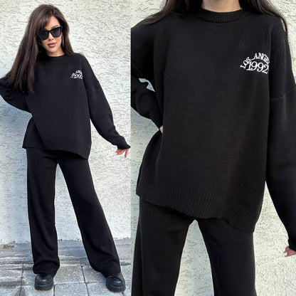 Woman wearing a black sweater and pants set with a brand logo, standing against a light-colored wall.