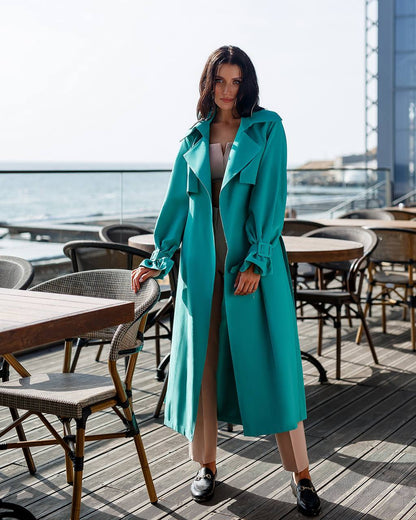 Woman in a turquoise coat standing on a rooftop with tables and chairs below.