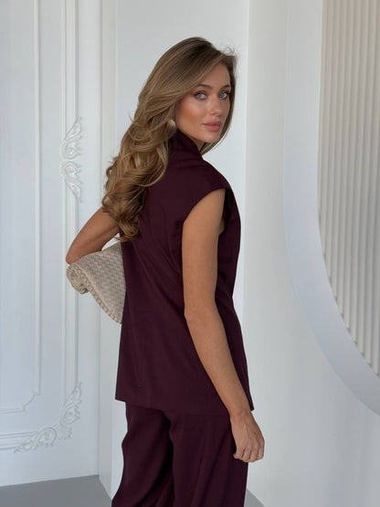 Woman in a burgundy sleeveless top and pants standing against a white wall.