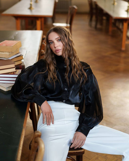 Woman sitting in a room with tables and books, wearing a black blouse and white pants.