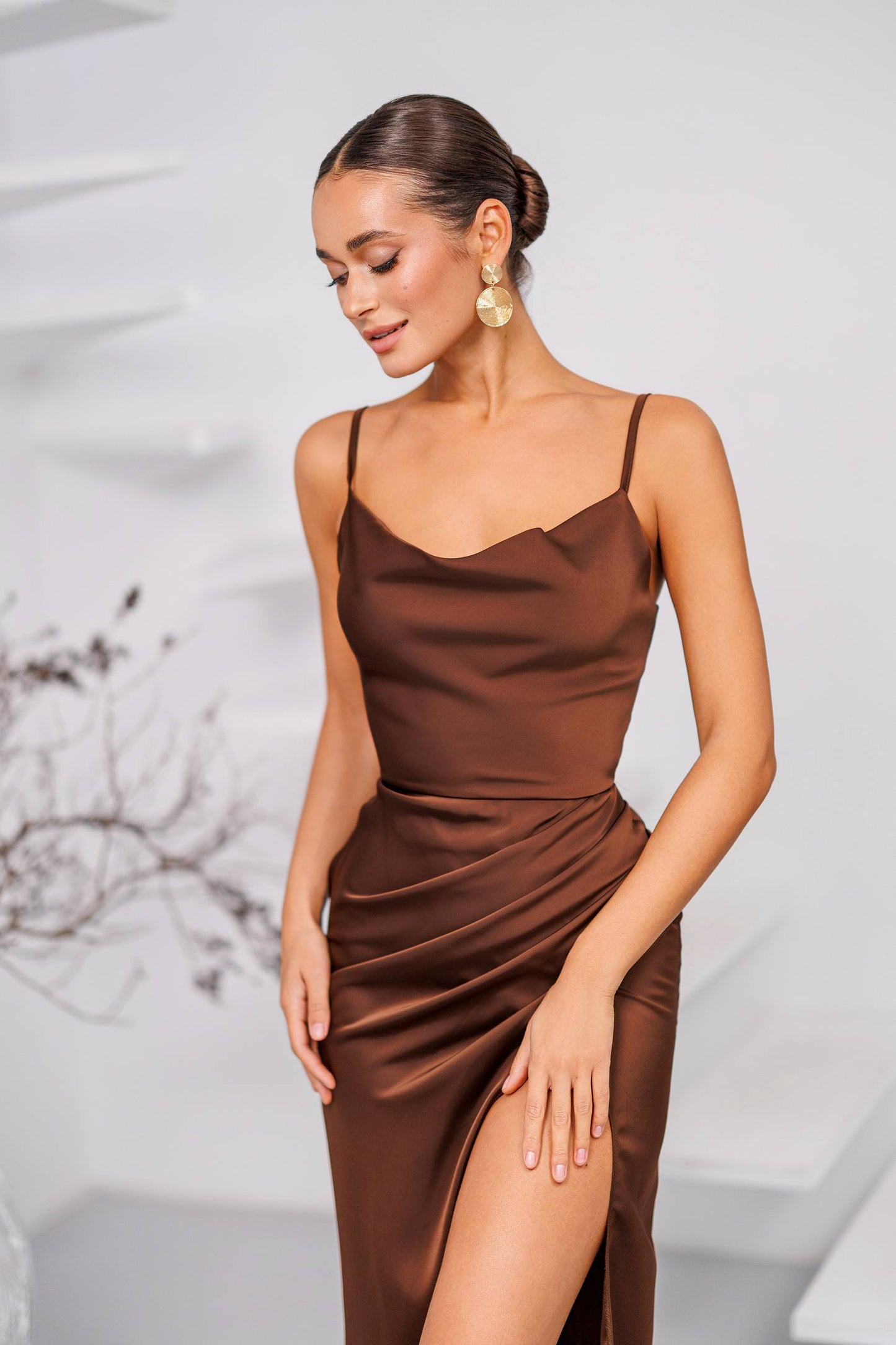 A woman wearing a brown satin slip dress with thin spaghetti straps, a sweetheart neckline, and a high thigh slit, posed against a light gray background.