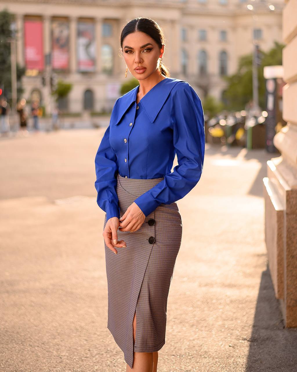 Woman wearing a blue blouse and gray skirt standing on a street with buildings in the background