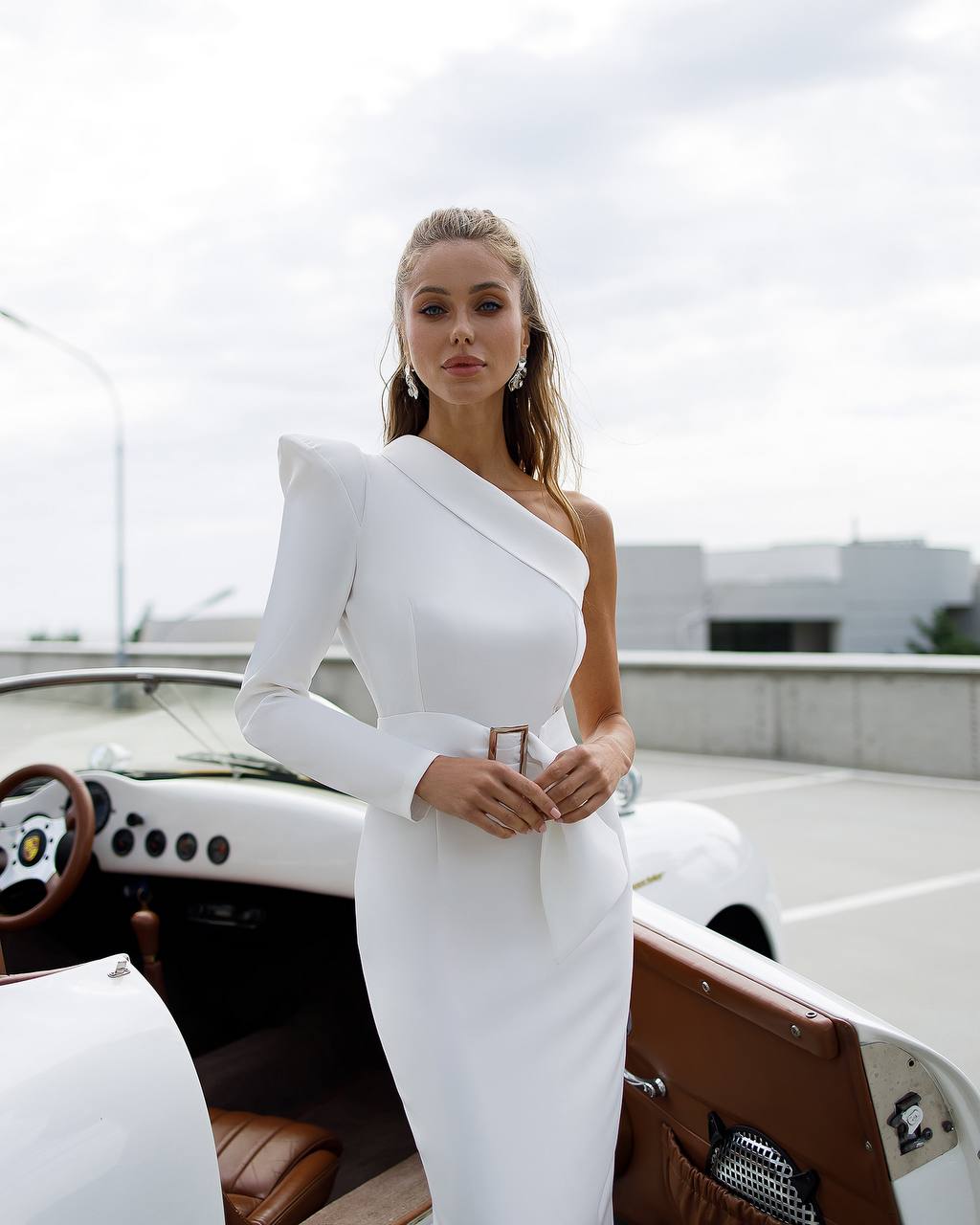 Woman in a white one-shoulder dress standing next to a vintage car on a rooftop.