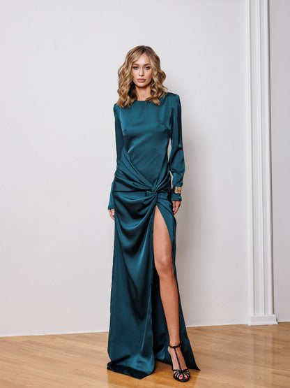 Woman wearing a teal satin dress with a high slit against a white wall.