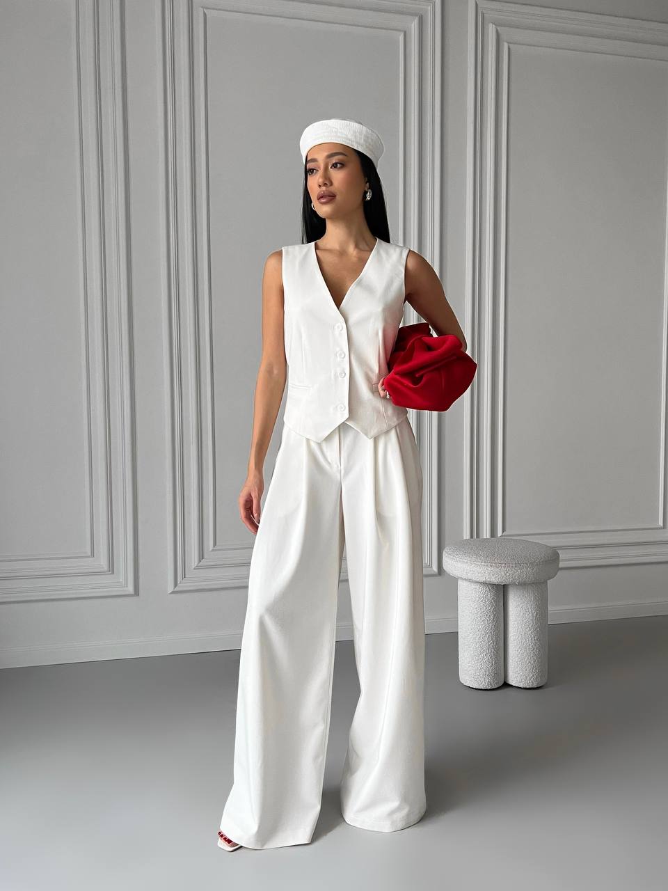 Woman in a white outfit holding a red clutch against a gray wall.
