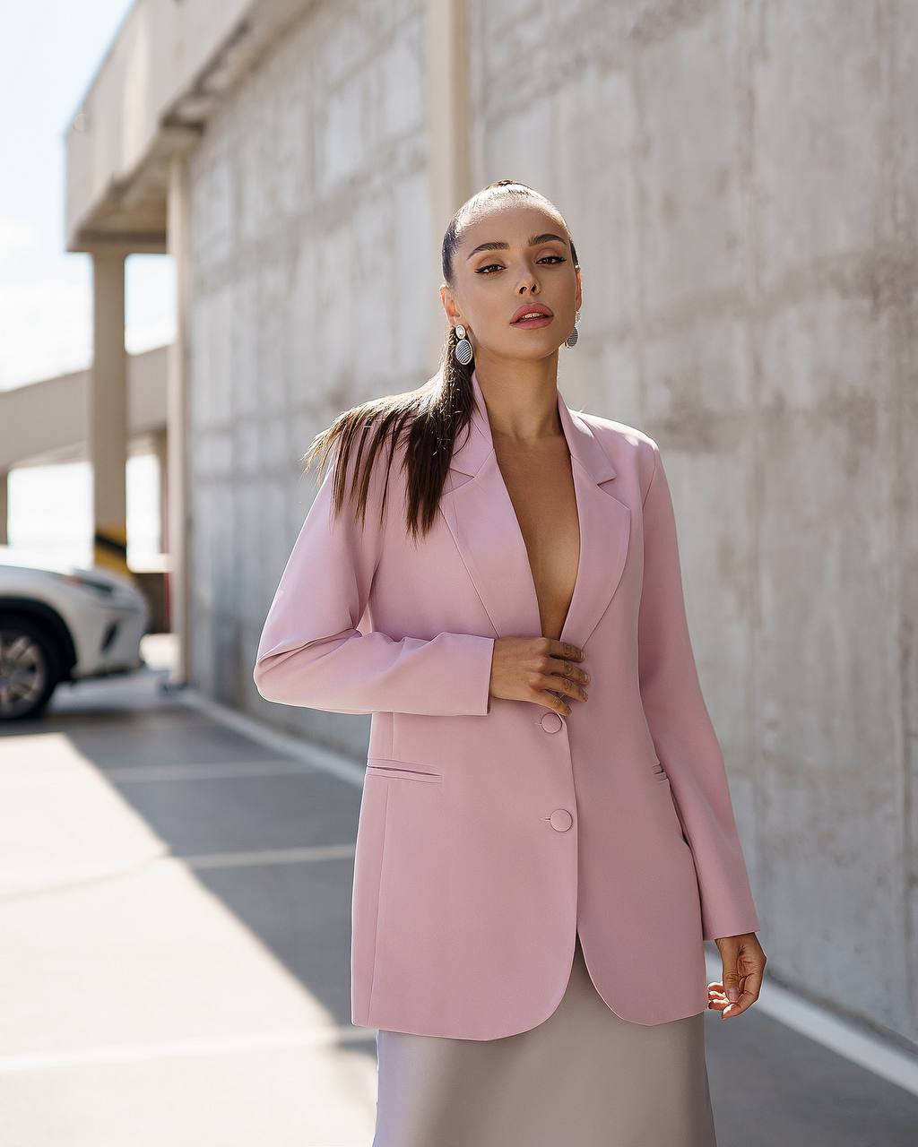 A woman wearing a light pink tailored blazer and a light gray skirt, standing in a concrete parking structure.