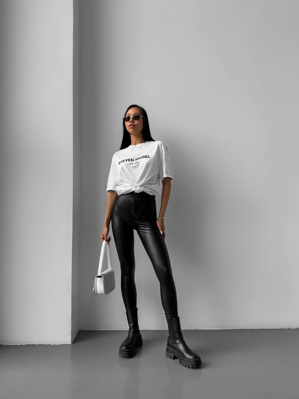 Woman wearing a white t-shirt, black leather pants, and black boots against a gray wall.
