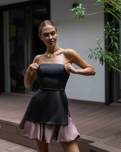 A woman models a strapless black peplum-style dress with a pleated light pink skirt, standing on a wooden deck in front of a modern house.