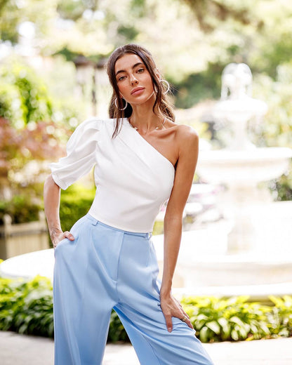 Woman wearing a white one-shoulder top and light blue pants outdoors.