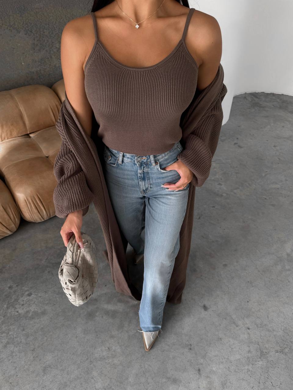 Person wearing a brown ribbed tank top and jeans, holding a beige purse in a minimalistic room.