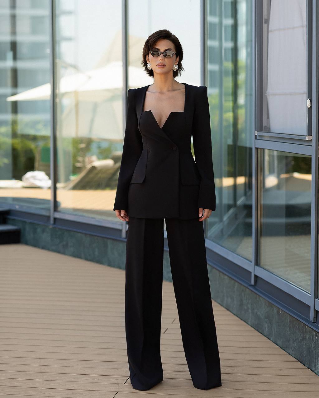 A woman stands on a sunlit balcony wearing a black tailored jumpsuit with structured shoulders, a deep V-neckline, and wide-leg pants. Large windows and outdoor seating are visible in the background.