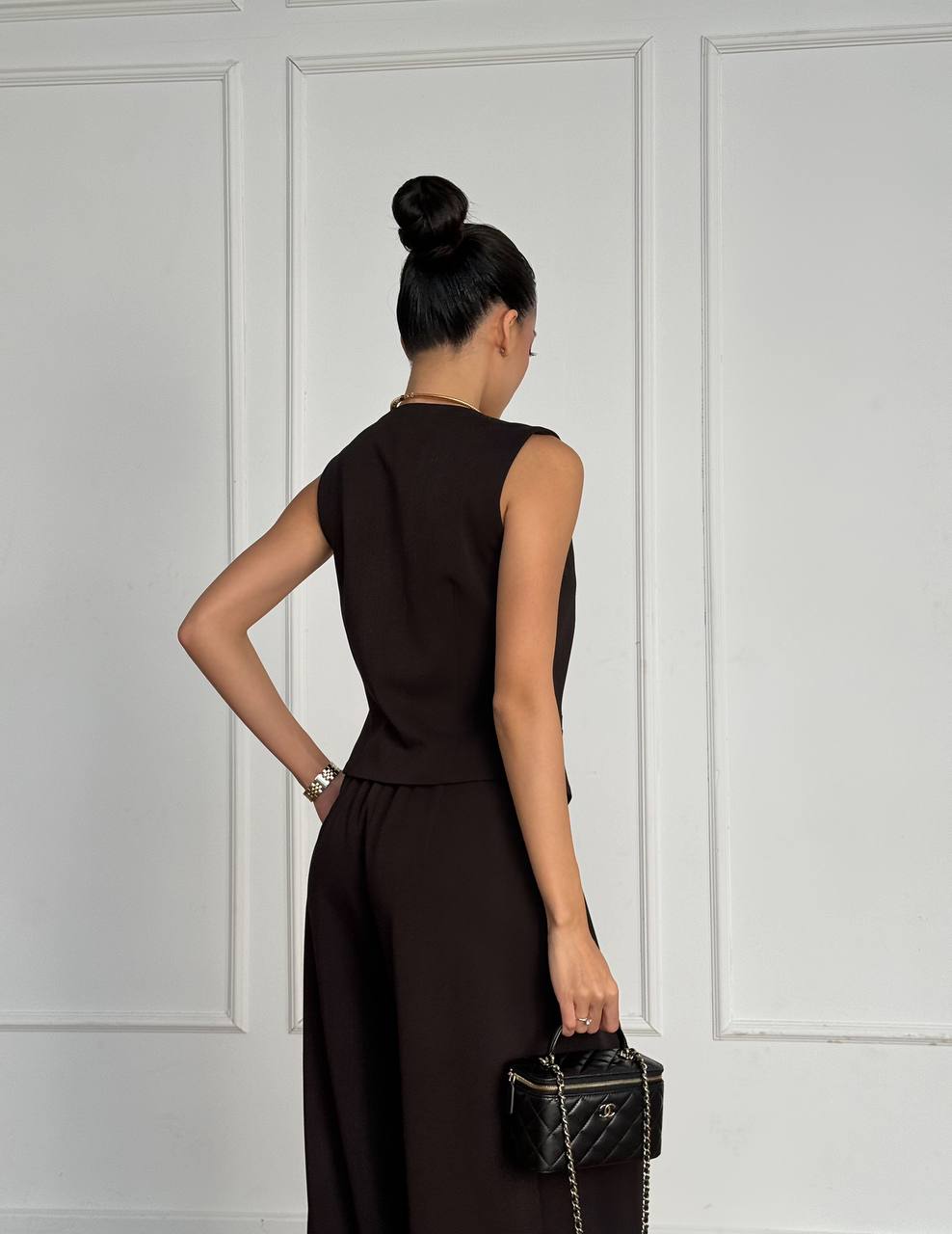 Woman in a brown dress holding a black handbag against a white paneled wall.