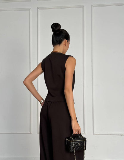 Woman in a brown dress holding a black handbag against a white paneled wall.